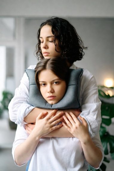 Young woman standing behind a child, holding a gray neck pillow around the child's neck, both with serious expressions in a light-filled room with plants in the background, symbolizing overprotective enabling that hinders growth.